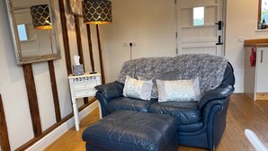 A living room with a dark leather sofa, a matching ottoman, a white side table with a decorative mirror, and two patterned lamps. - Gosfield Cartlodge Retreat (Halstead)