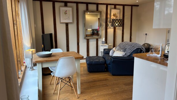 A modern living room with a wooden dining table, white chairs, a black leather sofa, and a wooden floor. - Gosfield Cartlodge Retreat (Halstead)