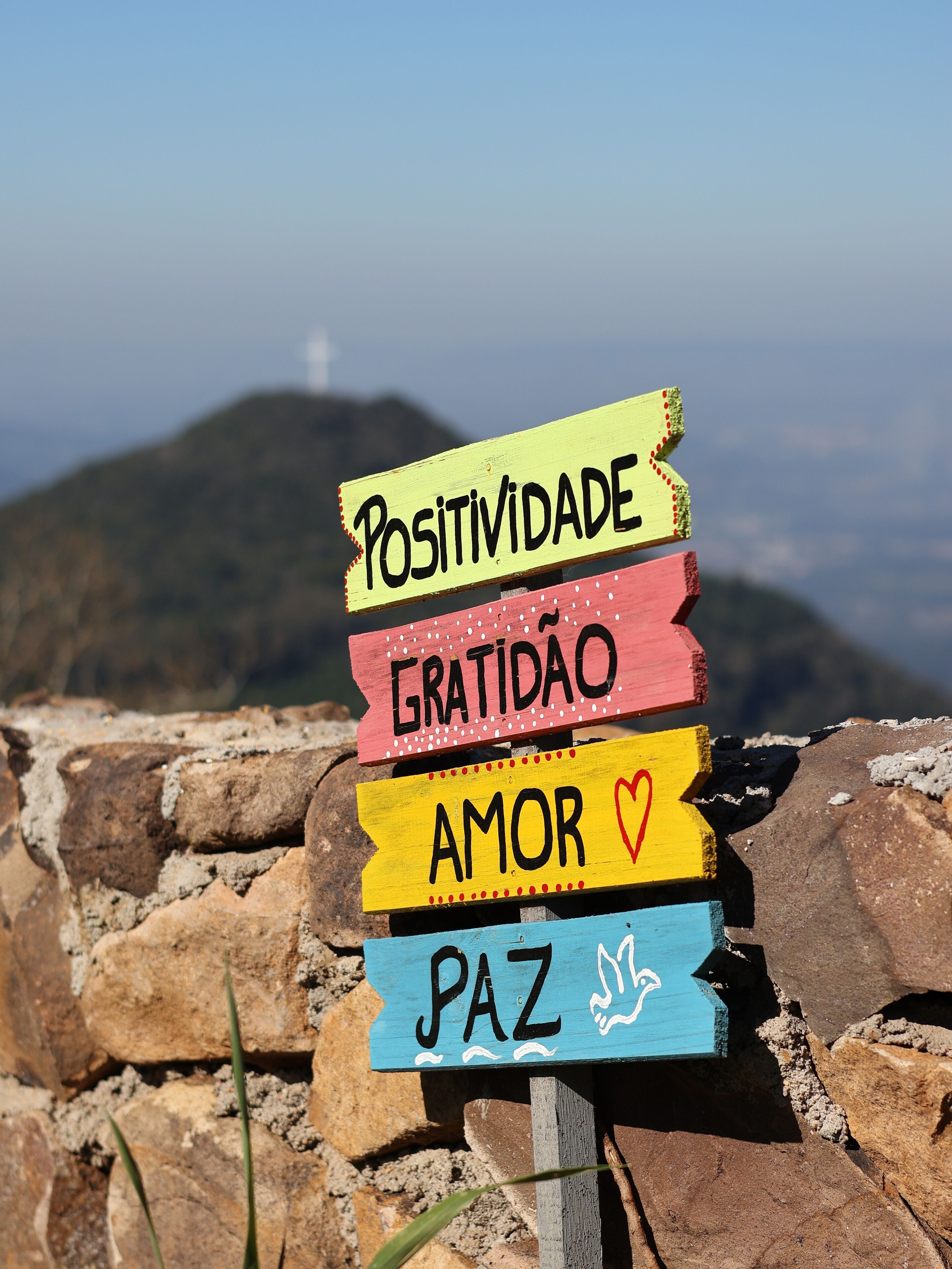 A wooden sign with words 'POSITIVIDADE,' 'GRATIDÃO,' 'AMOR,' and 'PAZ' on a mountain trail.