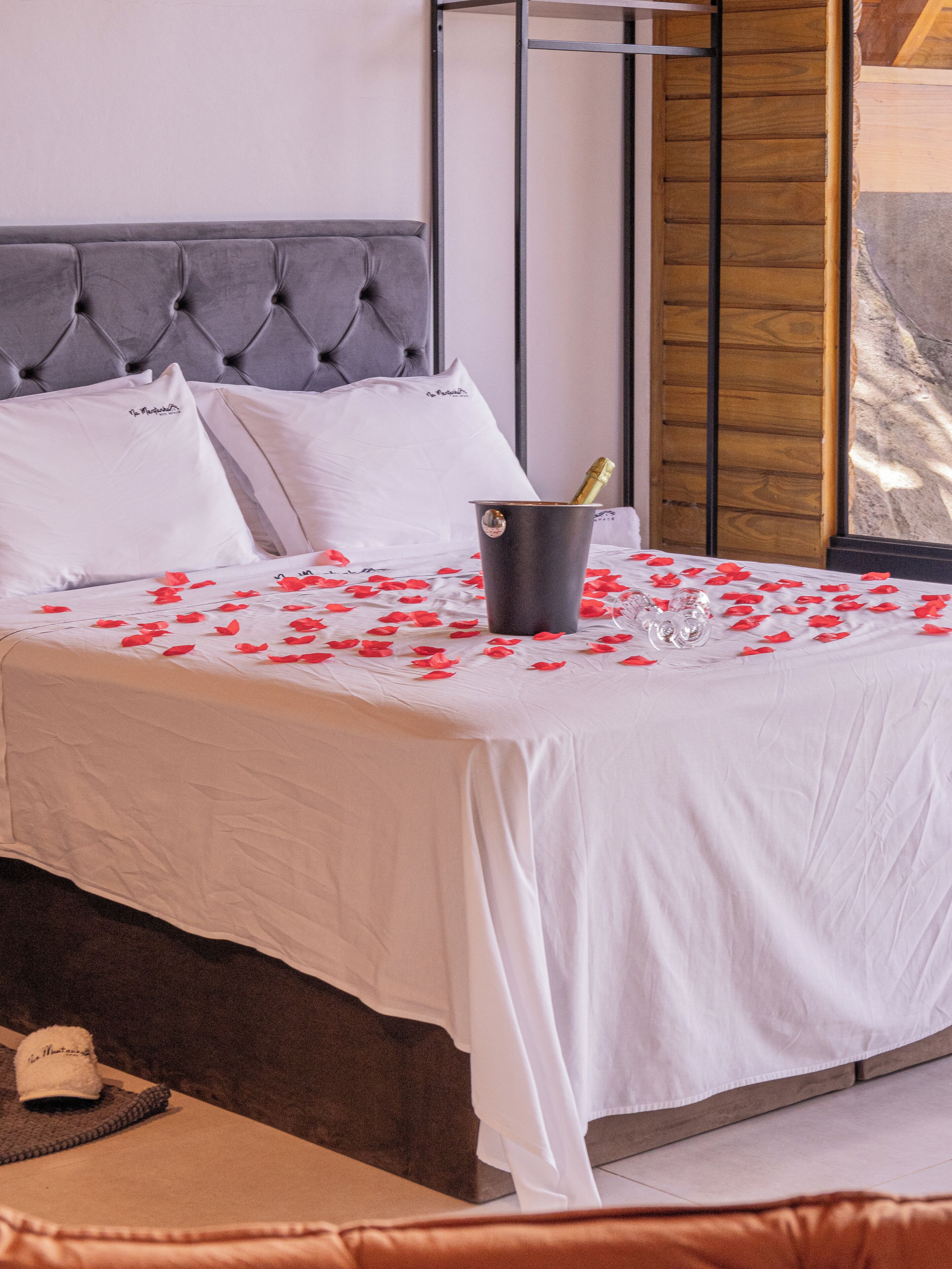 Una cama bien hecha con sábanas blancas, un balde con una botella de champán y pétalos de rosa esparcidos sobre la cama.