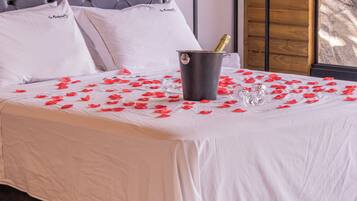 Una cama bien hecha con sábanas blancas, un balde con una botella de champán y pétalos de rosa esparcidos sobre la cama.