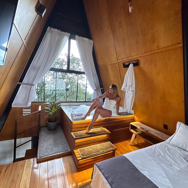 A cozy, modern bedroom with a wooden sloped ceiling, a large window with white curtains, a bed with a gray and white duvet, and a small bedside table with a towel.