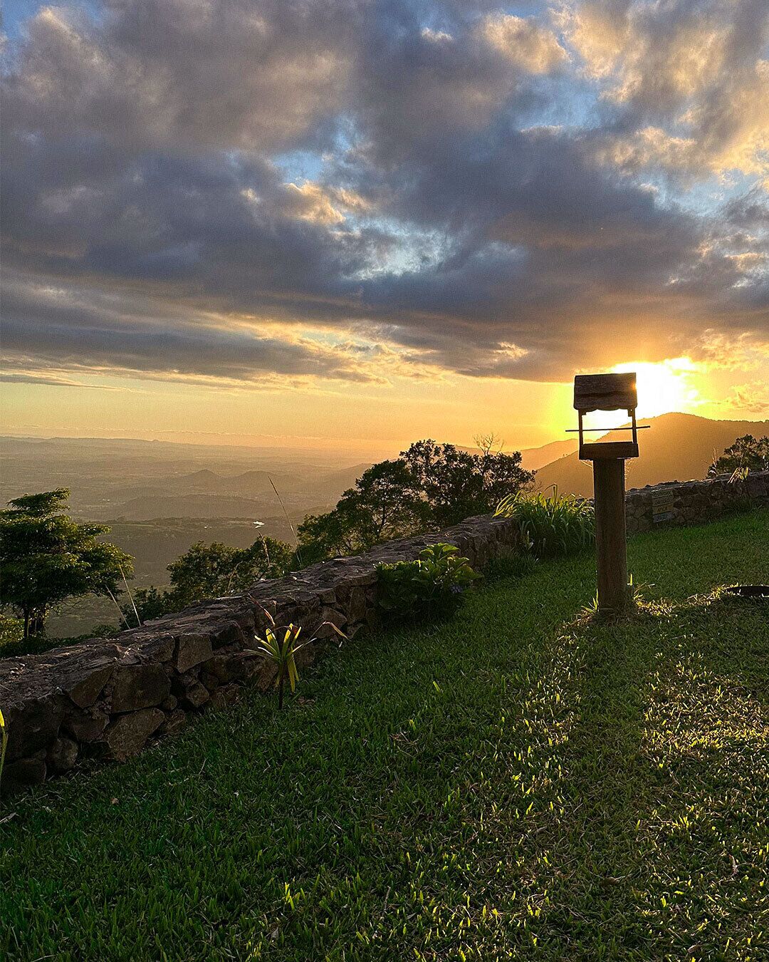A stone bench with a wooden sign, overlooking a grassy landscape with trees and a distant view of mountains under a cloudy sky.