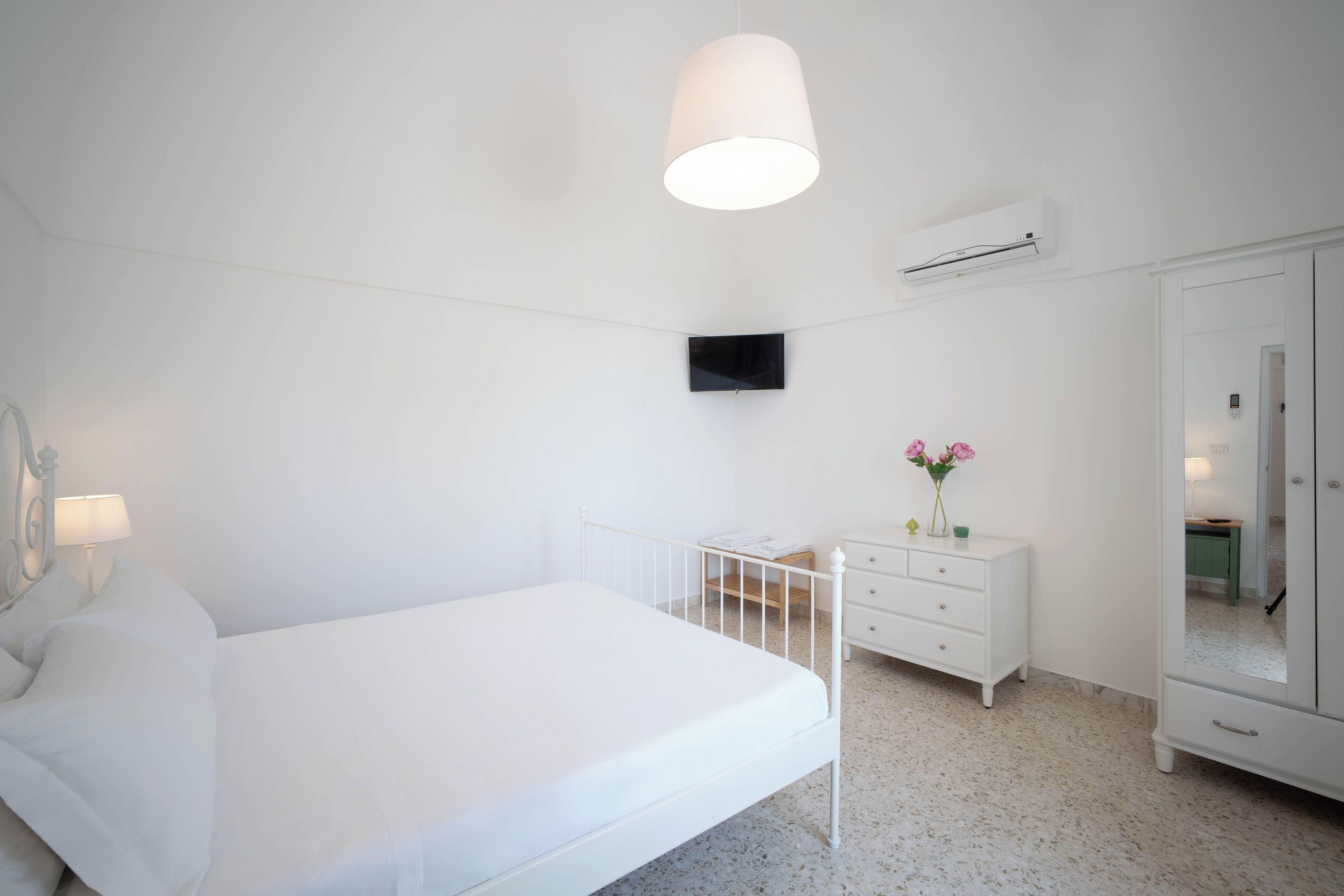 A white bedroom with a bed, a bedside table, a dresser, a mirror, and a wall-mounted TV.