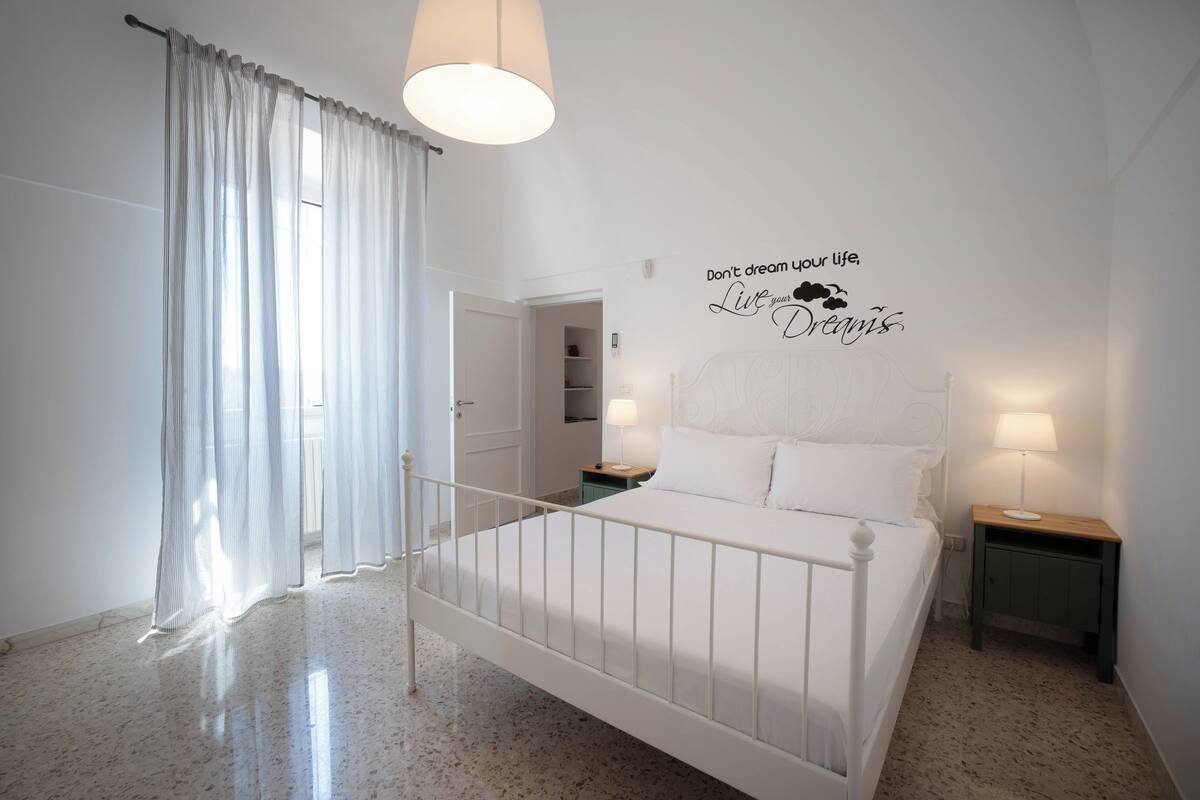 A bedroom with a bed, a nightstand, a lamp, a wall with a motivational quote, and a door leading to another room.