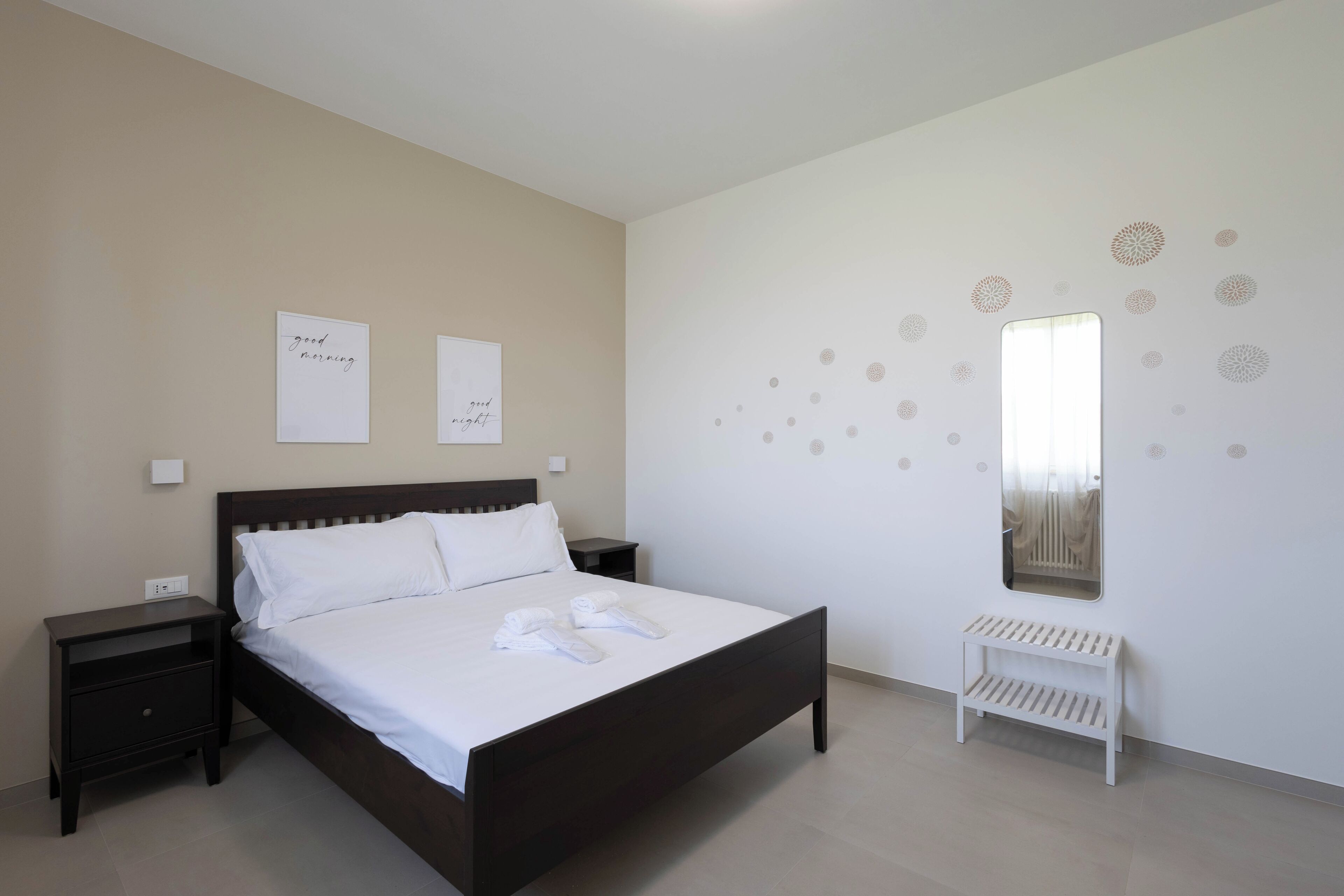 A bedroom with a dark wooden bed, white bedding, two bedside tables, a white bench, and decorative wall art.