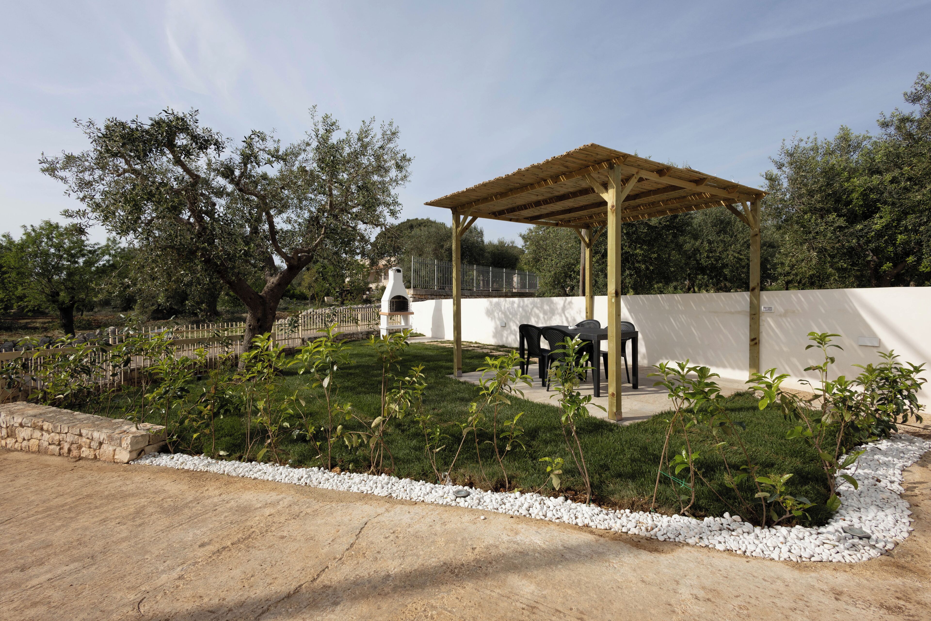 A covered outdoor seating area with a pergola, surrounded by a well-maintained garden with white gravel edging and various shrubs.