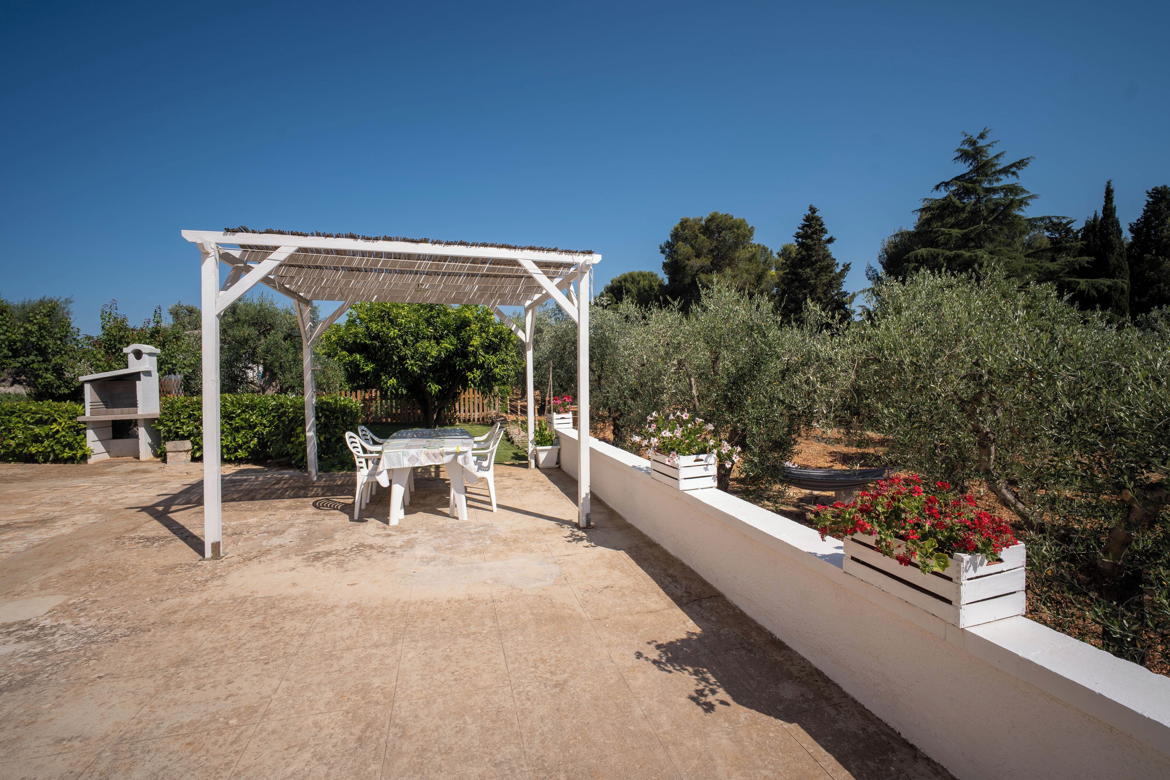 Un espace extérieur abrité, aménagé avec une table et des chaises, entouré de verdure et bordé d’un mur blanc.