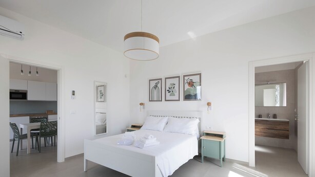 A modern bedroom with a white bed, bedside tables, and a hanging light fixture. There is a kitchen area visible through an open door, and a bathroom with a mirror and sink. - PORTA SUGLI ULIVI (Monopoli)