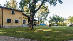 A yellow building with a stone base, a paved area with a tree, and a grassy lawn. - Villa Woodland Garden - Irpinia (Montemarano)