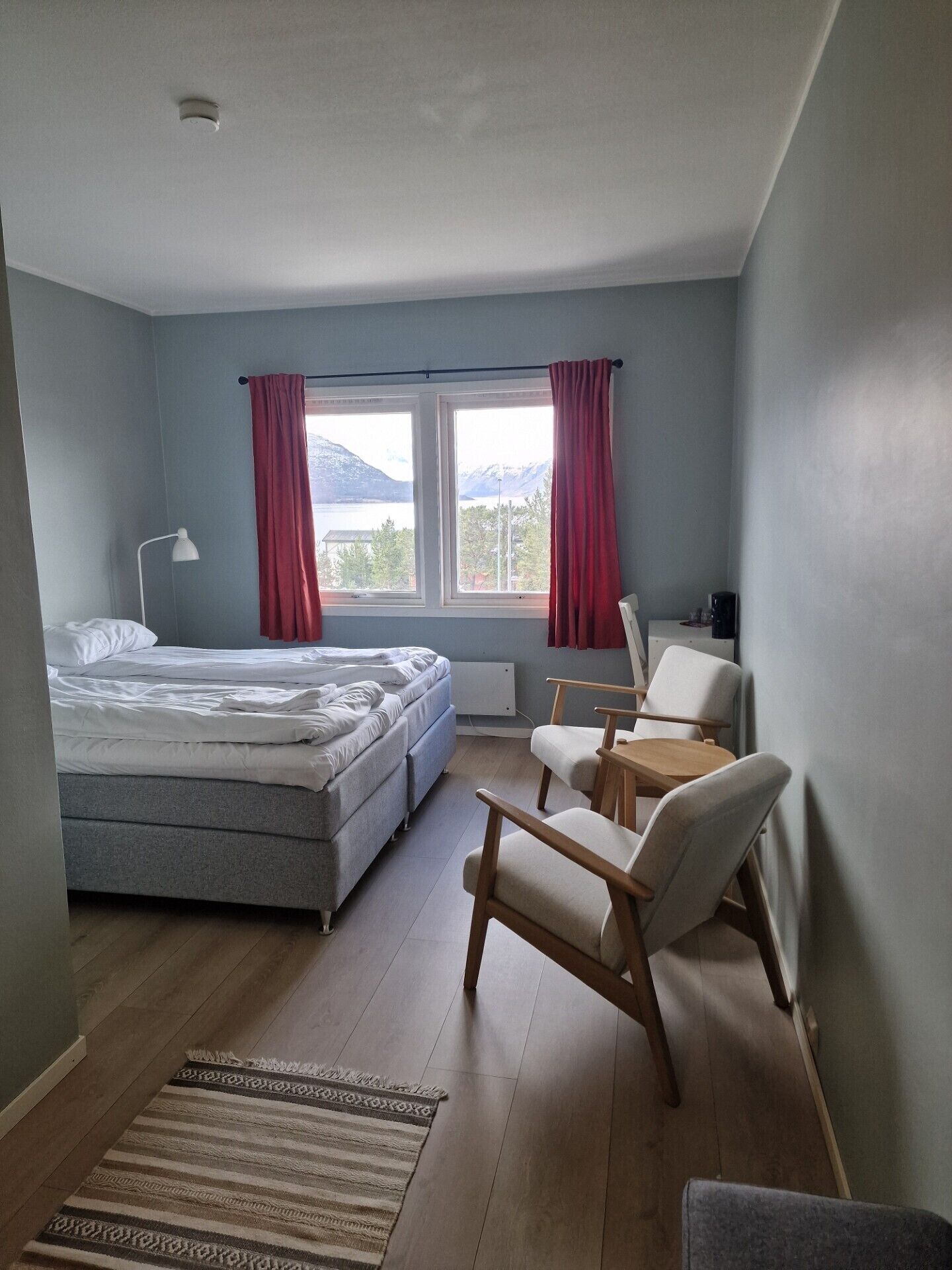 A bedroom with a bed, two chairs, a small table, a lamp, and a view of mountains through the window.