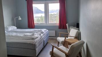 A bedroom with a bed, two chairs, a small table, a lamp, and a view of mountains through the window.