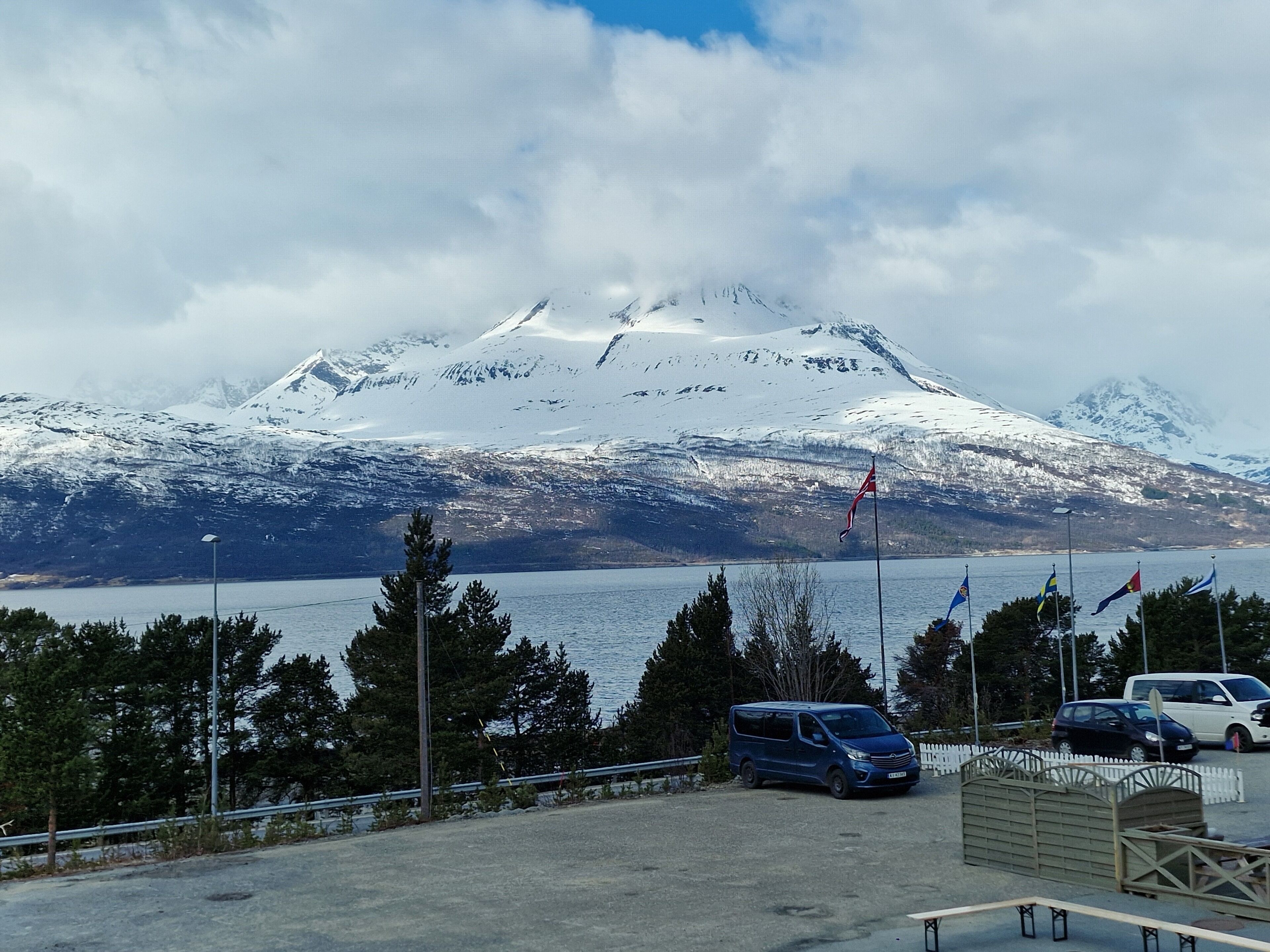 Un área de estacionamiento con vehículos, un asta con banderas y una montaña nevada al fondo.