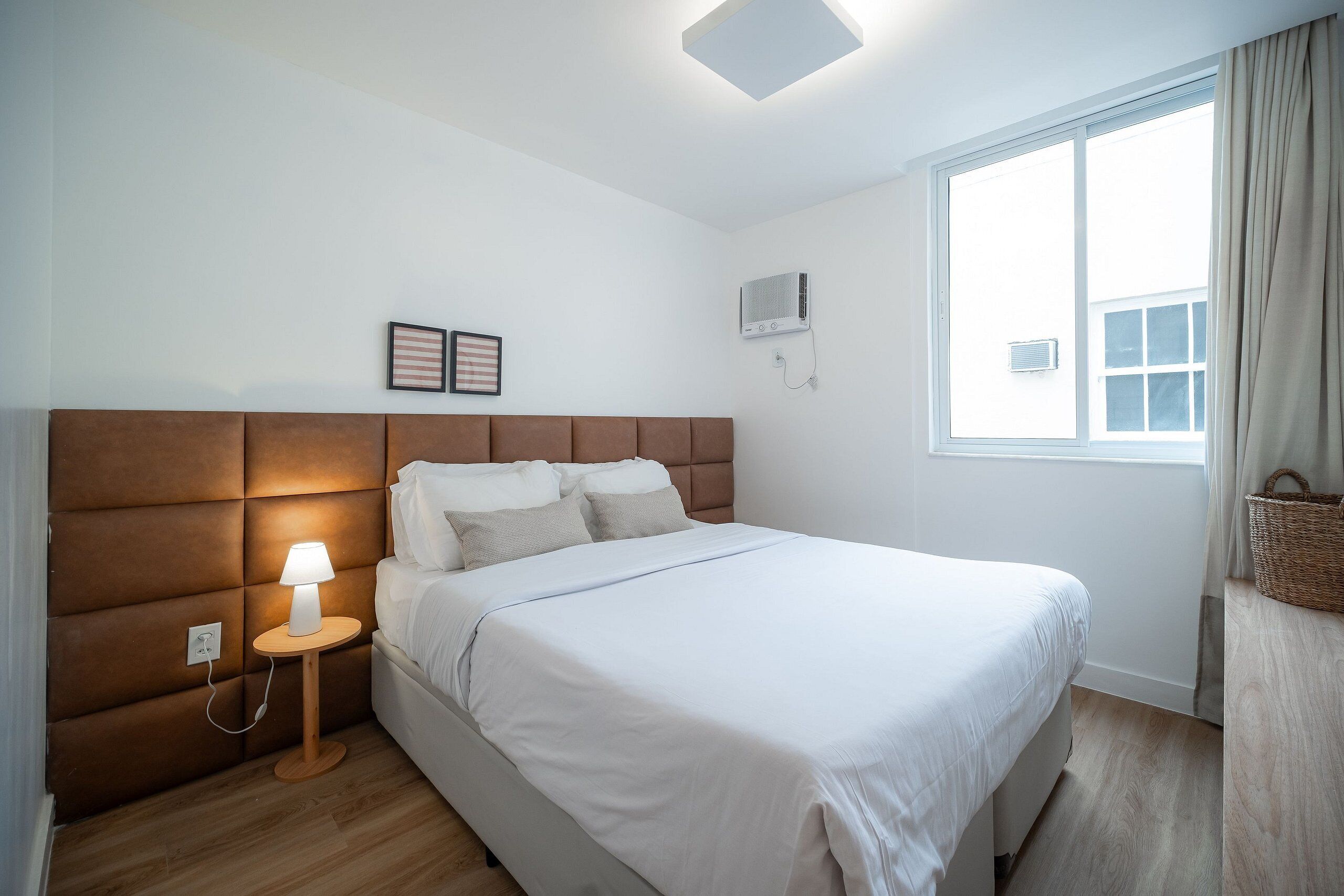A bedroom with a large bed, a bedside table with a lamp, a wall-mounted air conditioner, and a window with curtains.