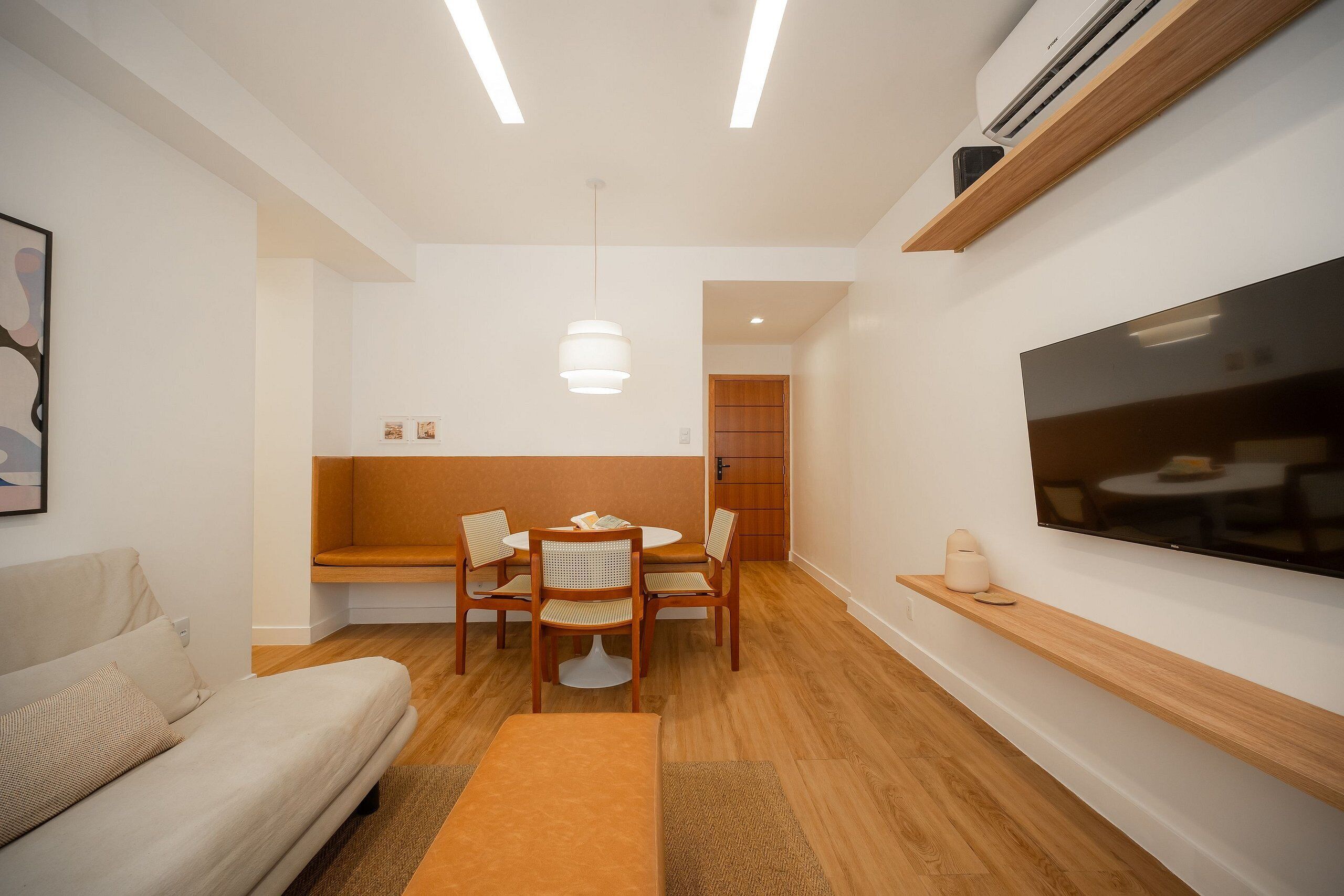 A modern living room with a sofa, a dining table with chairs, a TV mounted on the wall, and an air conditioning unit.