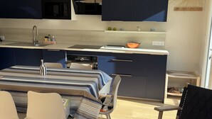 A modern kitchen with dark cabinetry, a microwave, and a built-in oven. There is a dining area with a striped tablecloth and chairs, and a black chair with a woven seat. - RENTAL HOUSE 2 BEDROOMS IN THE CENTER OF PORNIC FOR 4 PEOPLE (Pornic)
