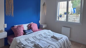 A bedroom with a blue accent wall, a bed with pink and white bedding, two hanging light fixtures, a window with a view of the outside, and a white bedside table. - RENTAL HOUSE 2 BEDROOMS IN THE CENTER OF PORNIC FOR 4 PEOPLE (Pornic)