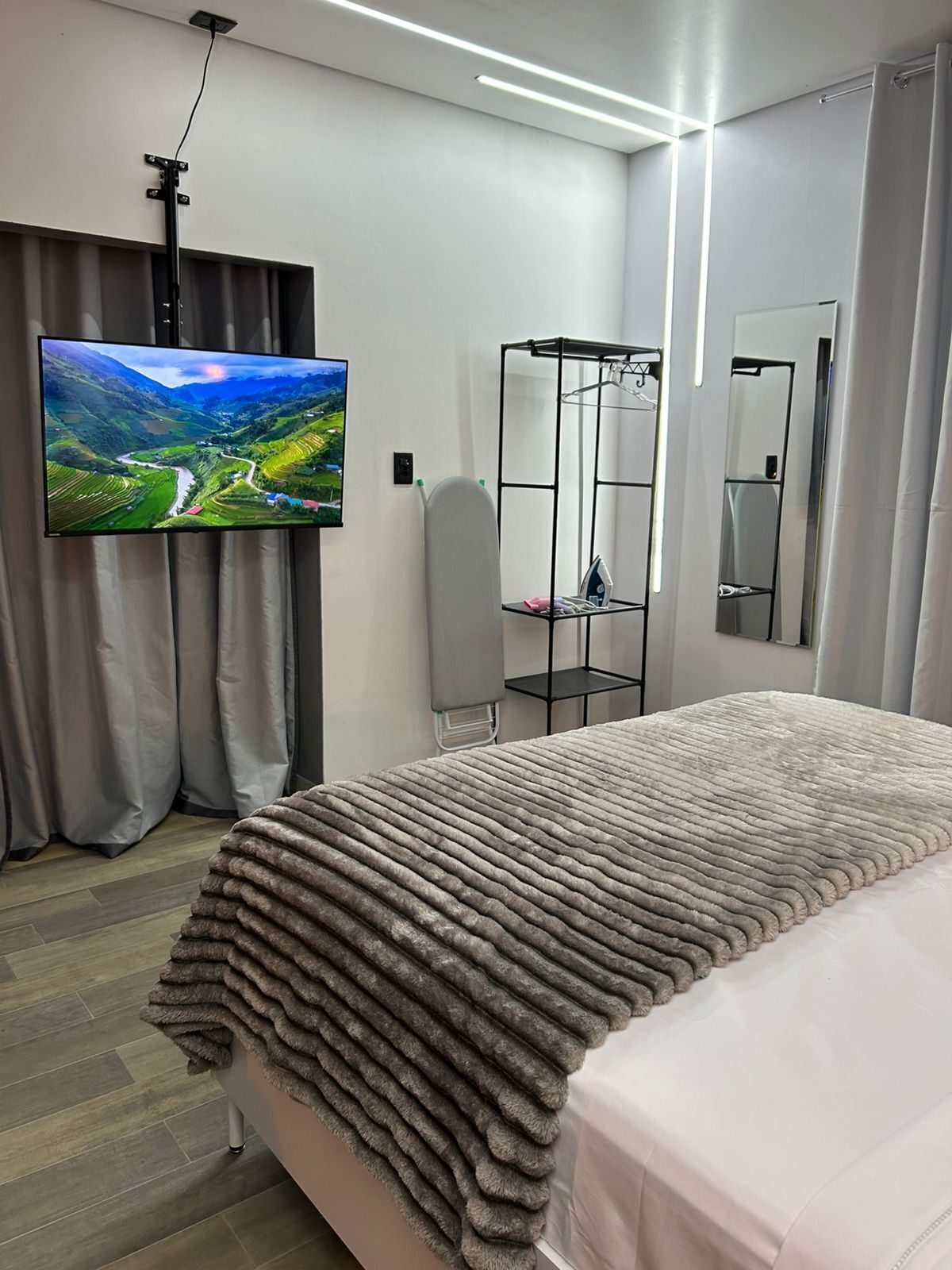 A modern hotel room with a large bed, a flat-screen TV mounted on the wall, a wardrobe with a mirror, and a small shelf with personal items.