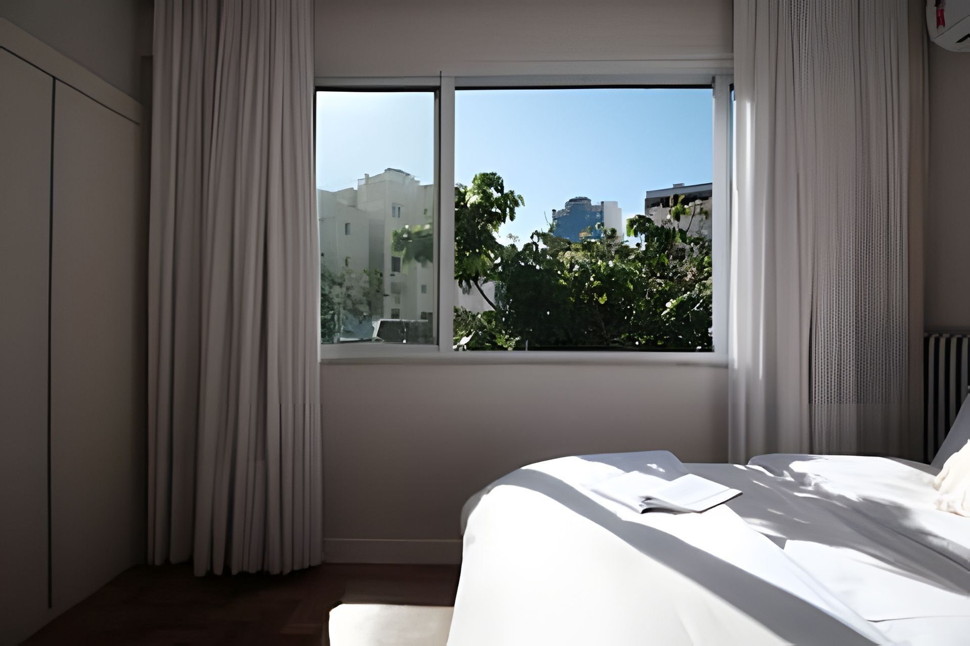 A bedroom with a large window, a bed, and a view of buildings and trees outside.