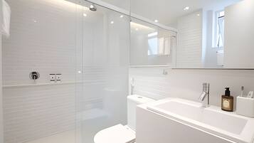 Uma casa de banho moderna com cabine de duche em vidro, sanita branca e lavatório branco com espelho em cima.