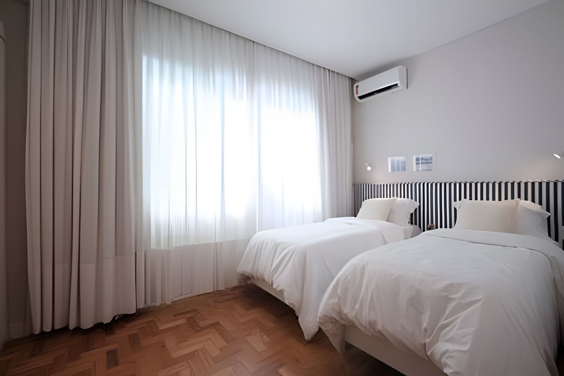 A hotel room with two beds, a large window with sheer curtains, a radiator, and a wall-mounted air conditioner.