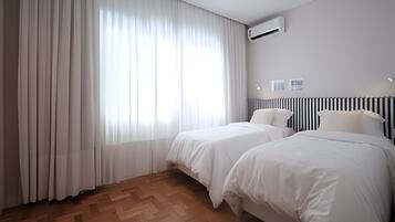 A hotel room with two beds, a large window with sheer curtains, a radiator, and a wall-mounted air conditioner.