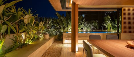 A modern outdoor dining area with a pool, wooden flooring, and tropical plants.