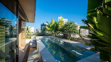Uma piscina no terraço com água azul cristalina, rodeada por vegetação exuberante e edifícios modernos ao fundo.