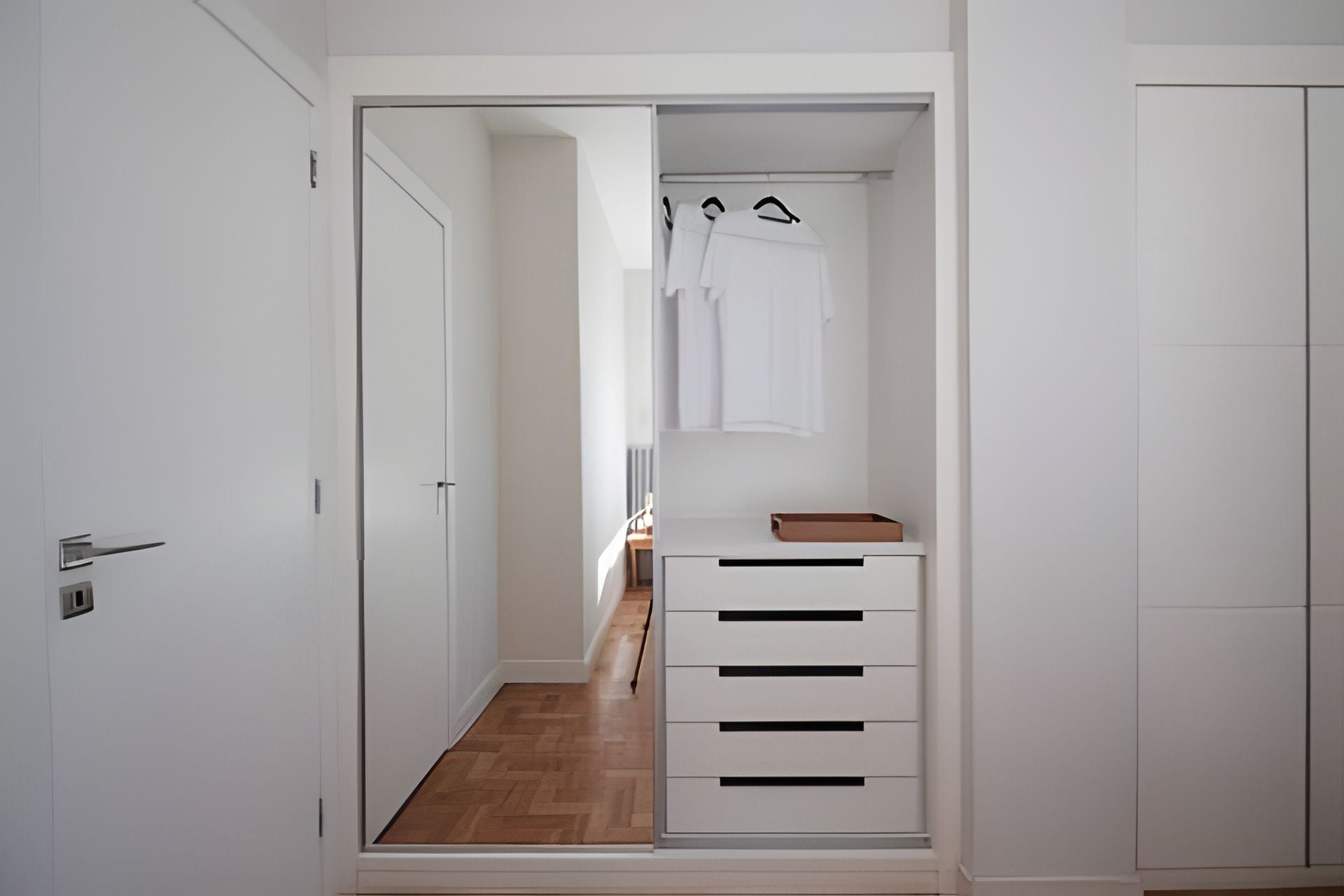 Um corredor estreito com um guarda-roupa branco, soalho de madeira e uma pequena mesa com um tabuleiro.