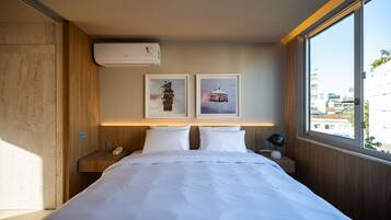 A hotel room with a bed, two wall-mounted air conditioning units, a bedside table with a lamp, and two framed pictures on the wall.