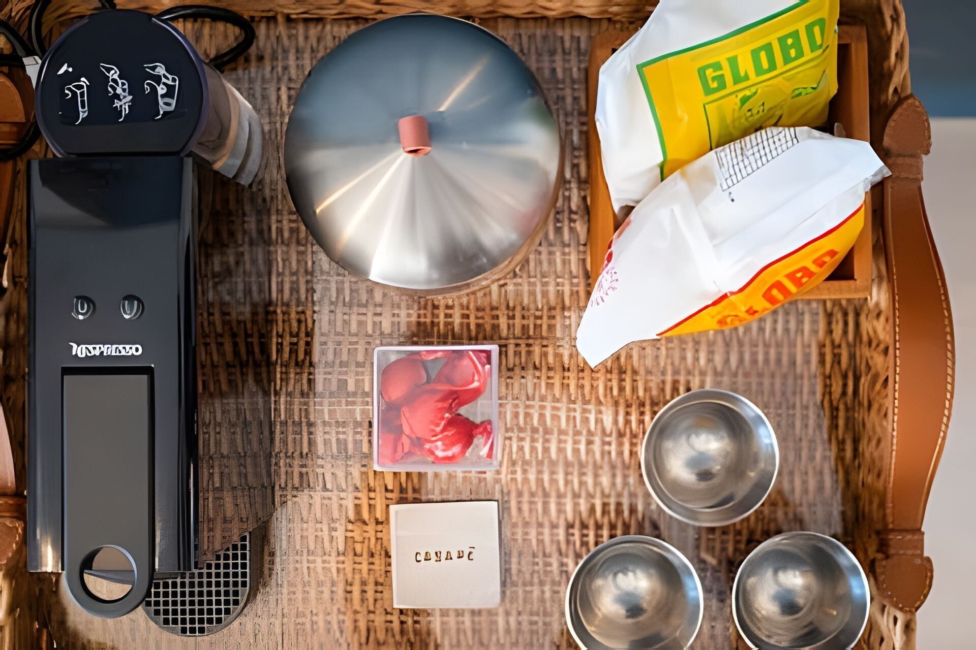Uma bandeja com um aparelho preto, uma chaleira prateada, uma bolsa amarela identificada com a etiqueta 'GLOBO', um objeto vermelho e três tigelas de metal.