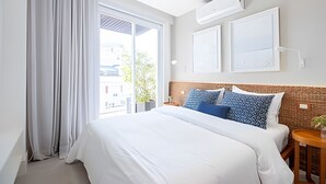 A hotel room with a large bed, a window with curtains, an air conditioning unit, and a small table with a plant.