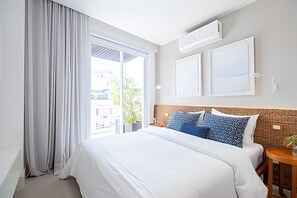 A hotel room with a large bed, a window with curtains, an air conditioning unit, and a small table with a plant. - Rua Dias Ferreira, 646 - Tesouro Escondido no Leblon (Rio de Janeiro)