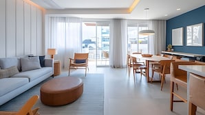 A modern hotel room with a sofa, dining table, chairs, and a large window offering a city view.