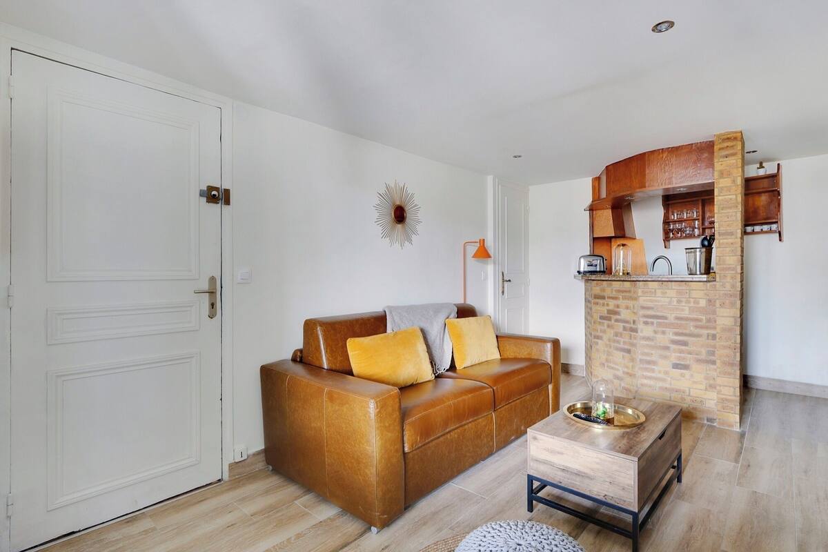 Un salon avec un canapé en cuir marron, une table basse en bois et un coin cuisine doté d’un mur de fond en briques et de meubles de rangement en bois.