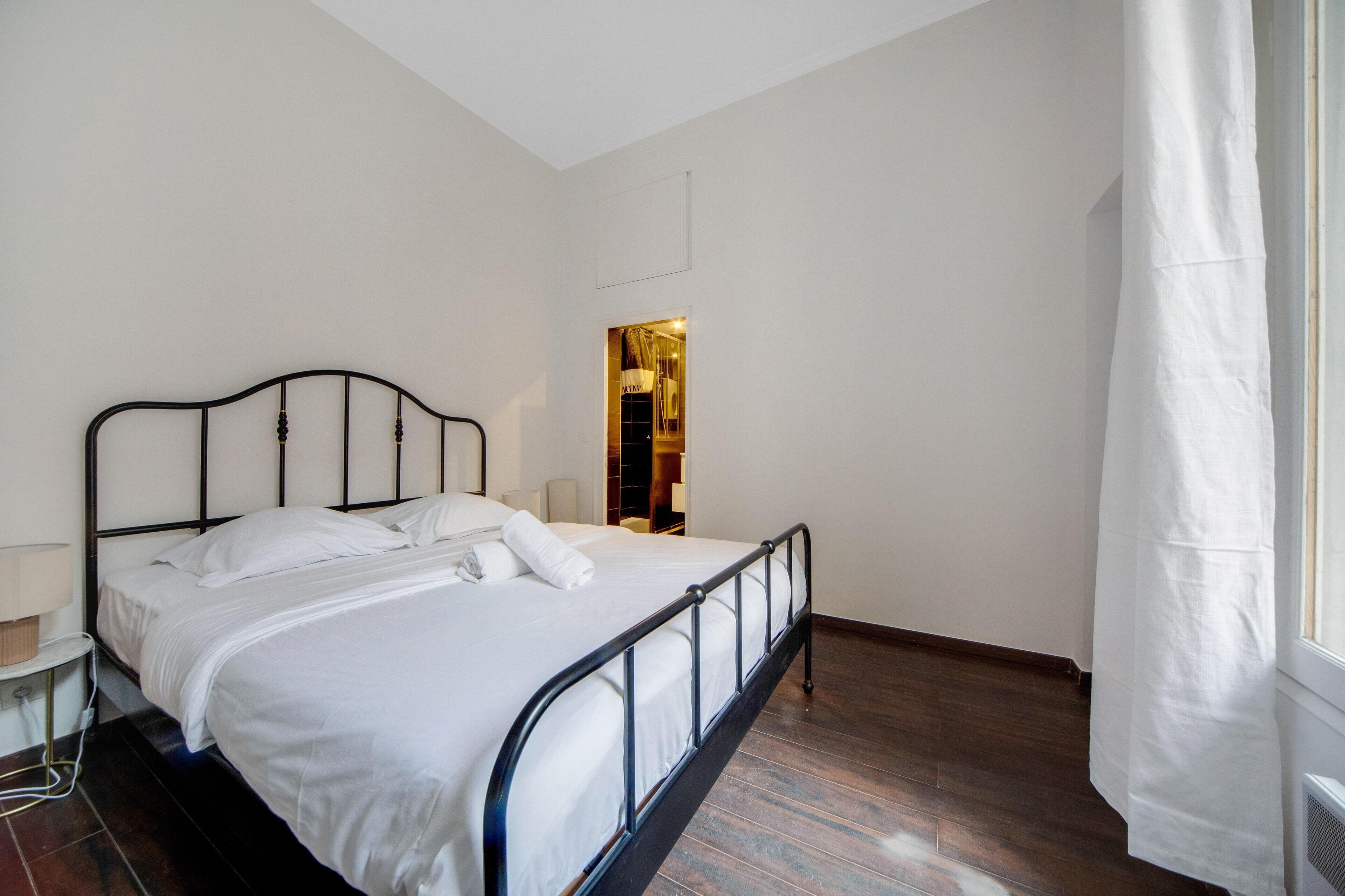 A neatly made bed with white linens in a room with wooden flooring and a window with white curtains.