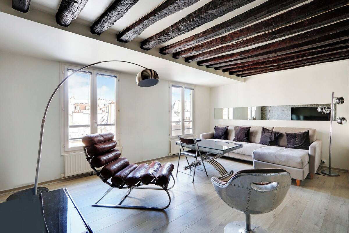 Un salon moderne avec un fauteuil en cuir, une table basse en verre, un canapé et une chaise en métal. De grandes fenêtres offrent une vue sur les bâtiments environnants.