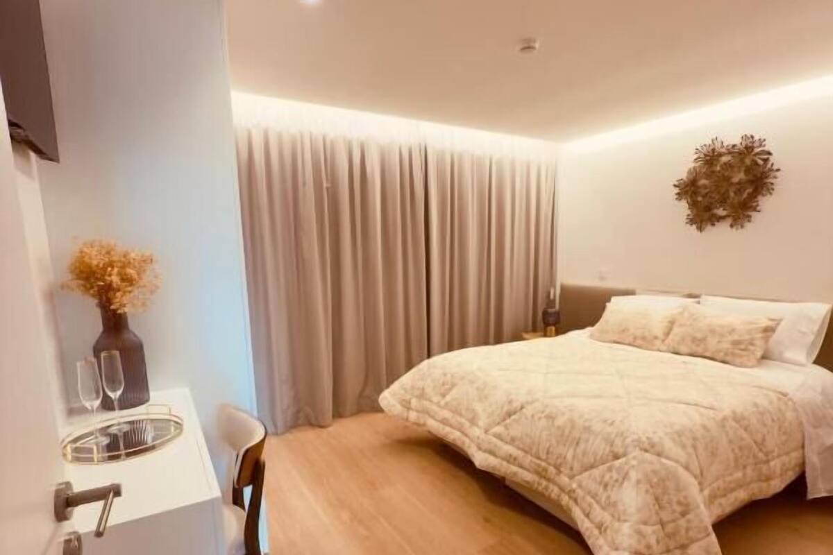 A modern bedroom with a large bed, a bedside table with a vase and glasses, and a wooden floor.