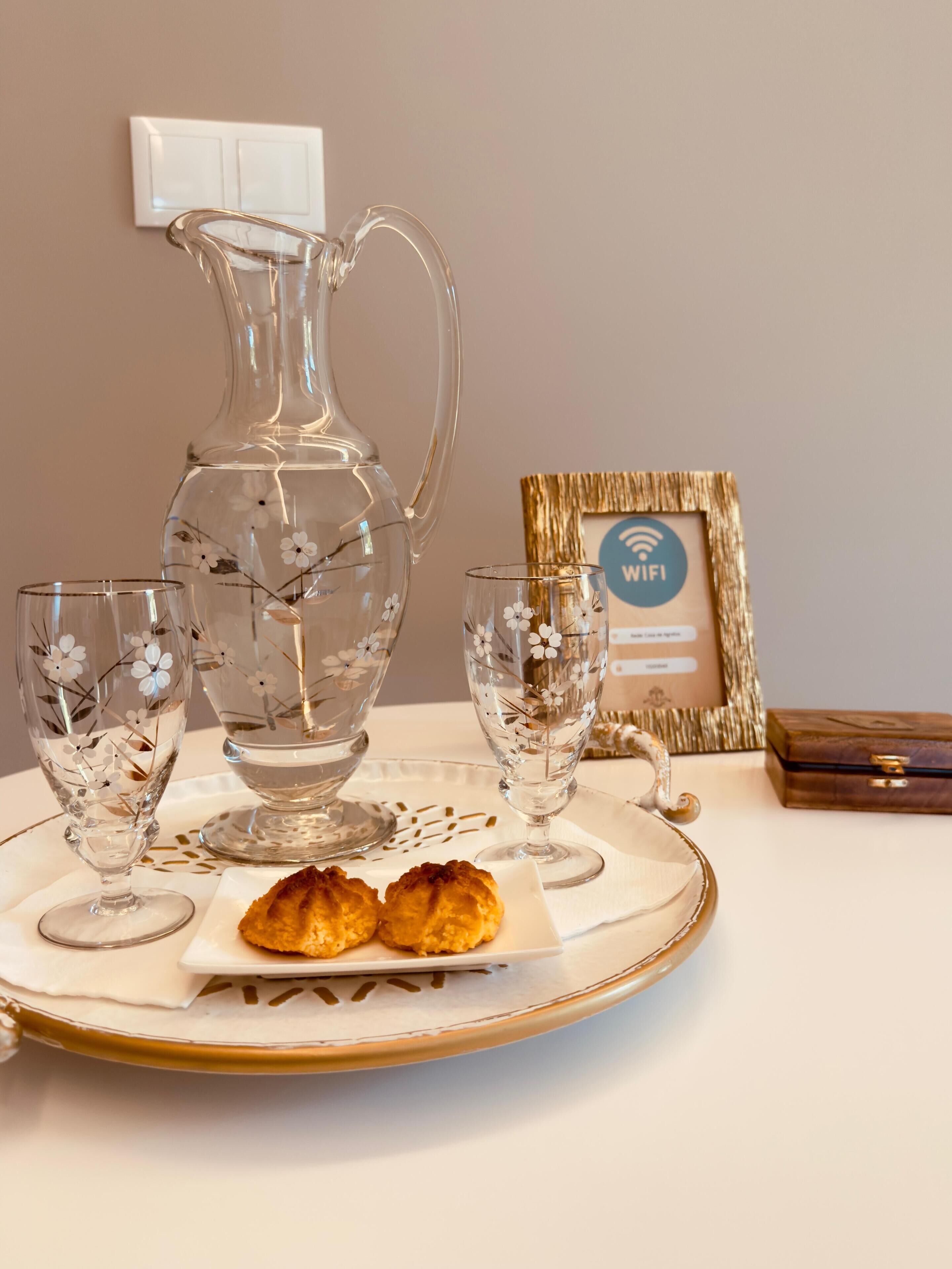 Uma mesa posta com uma jarra transparente, dois copos com desenhos florais e duas sobremesas em um prato.