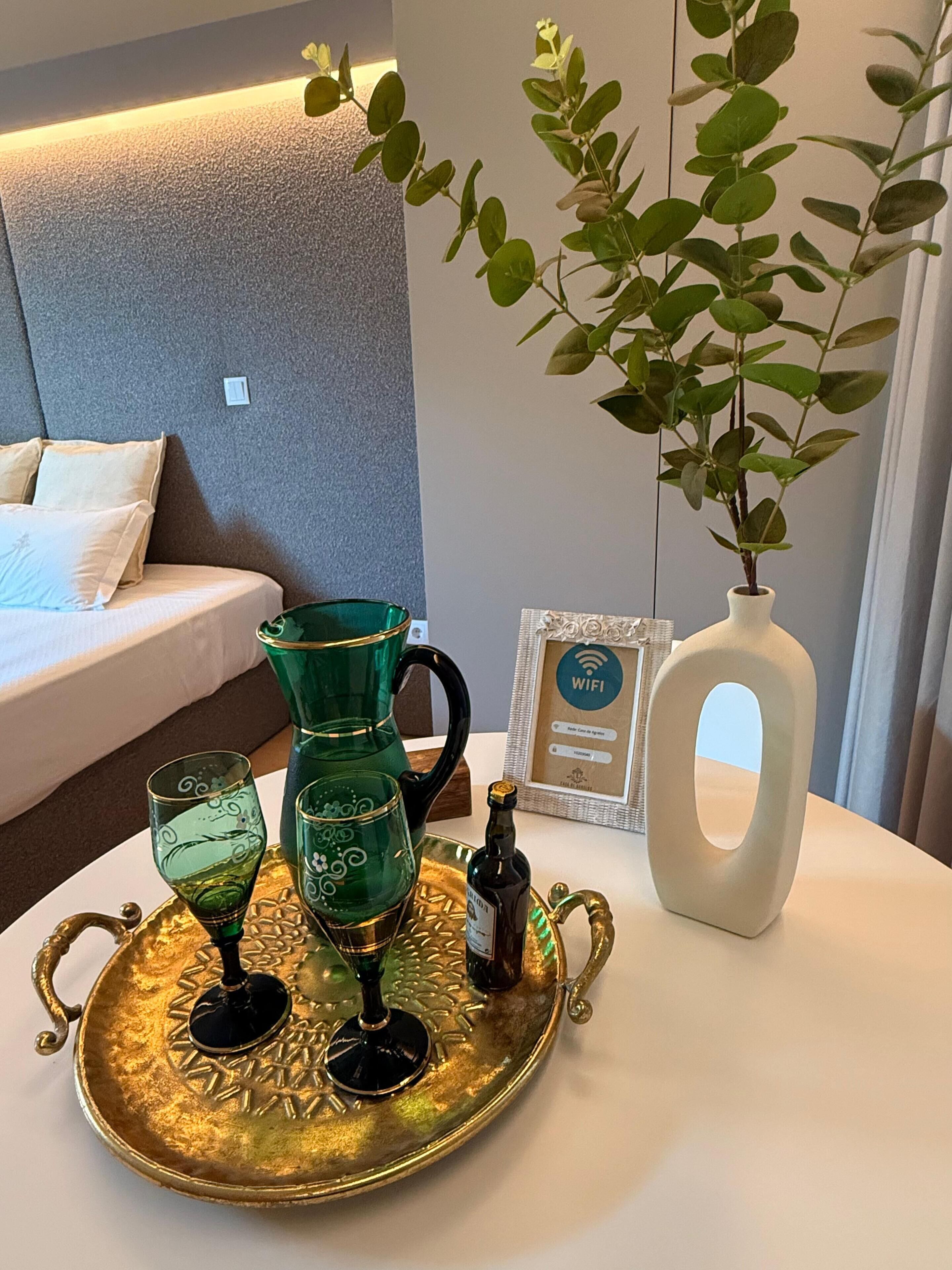 A hotel room with a bed, a bedside table with a tray holding glasses and a bottle, a vase with a plant, and a framed WiFi sign.