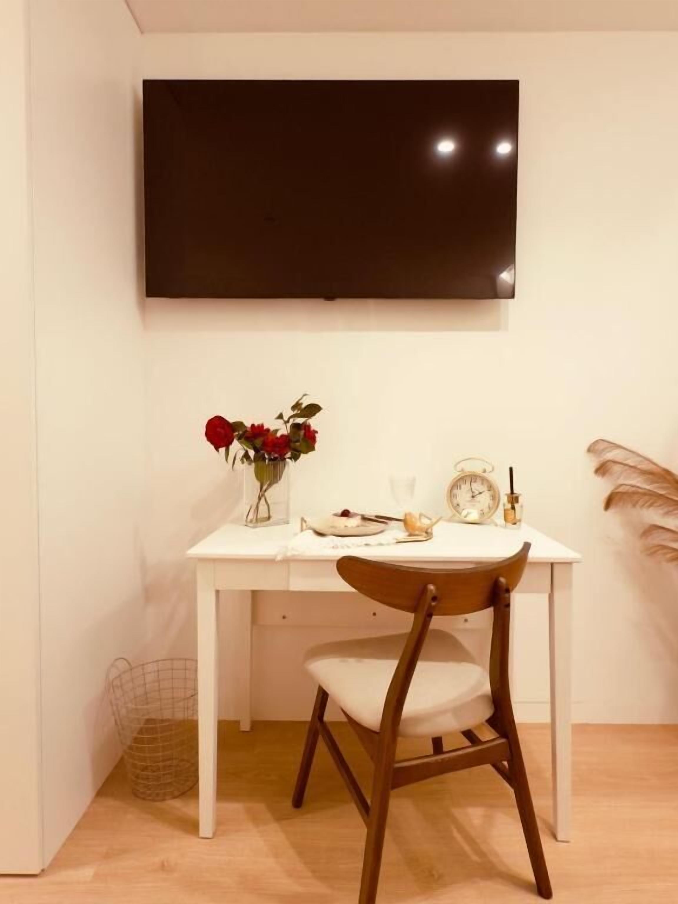 Un comedor con una mesa blanca, una silla de madera, un florero con flores rojas, un reloj y una planta pequeña.