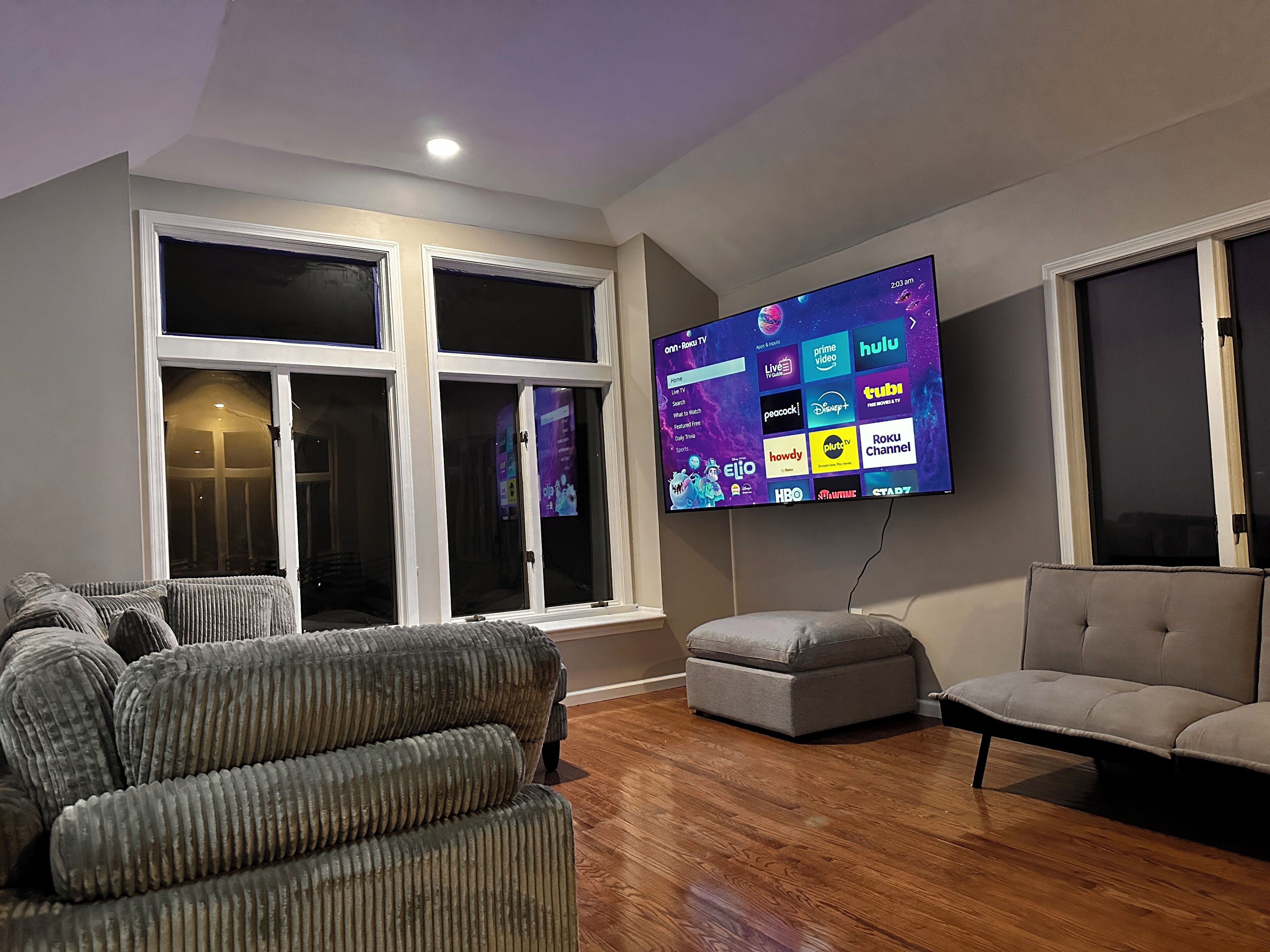 Una sala de estar con un televisor de pantalla plana montado en la pared, un sofá gris, un reposapiés gris y piso de madera.