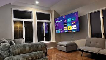 Una sala de estar con un televisor de pantalla plana montado en la pared, un sofá gris, un reposapiés gris y piso de madera.