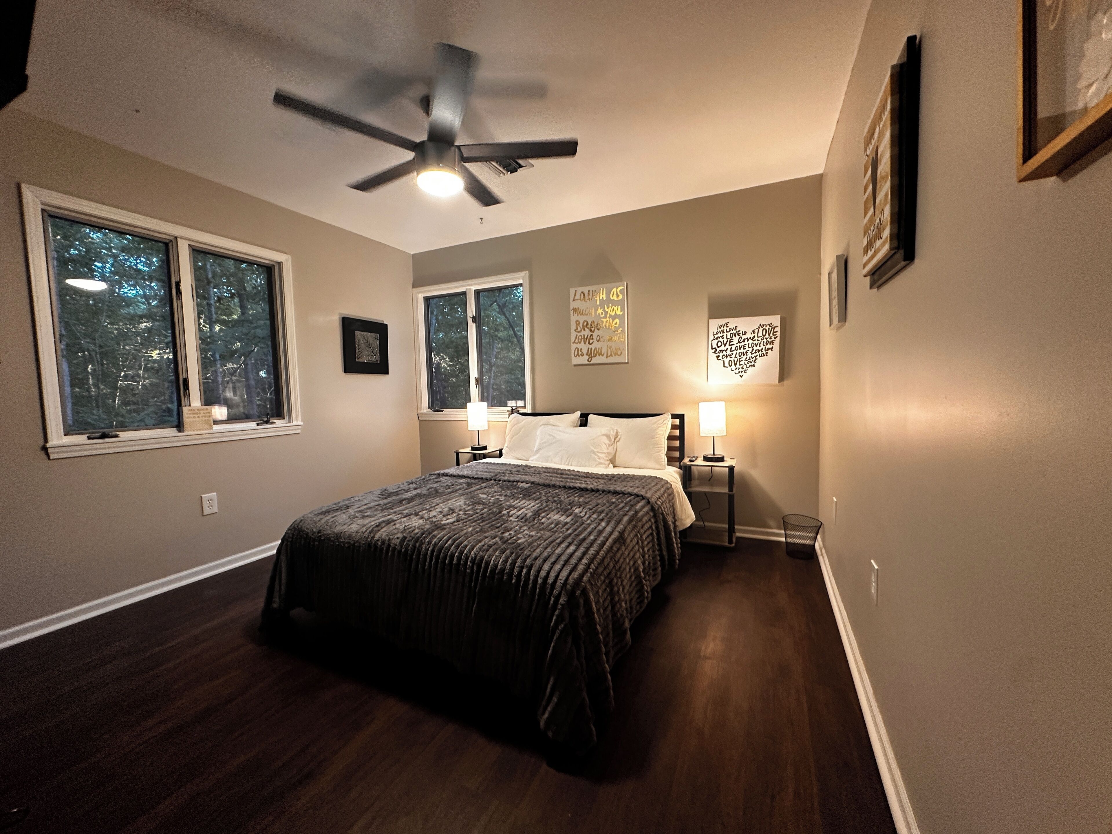 A bedroom with a bed, two bedside tables, a ceiling fan, and two windows.