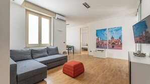 A modern living room with a grey sectional sofa, a red ottoman, wooden flooring, a flat-screen TV, and a painting of a historic building. - Vetro Apartment by Wonderful Italy  (Palermo)