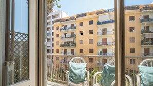 A balcony with wicker chairs and cushions, overlooking a row of yellow buildings with balconies. - Vetro Apartment by Wonderful Italy  (Palermo)