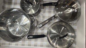 A kitchen drawer with four stainless steel pots and pans, each with a black handle and a lid.