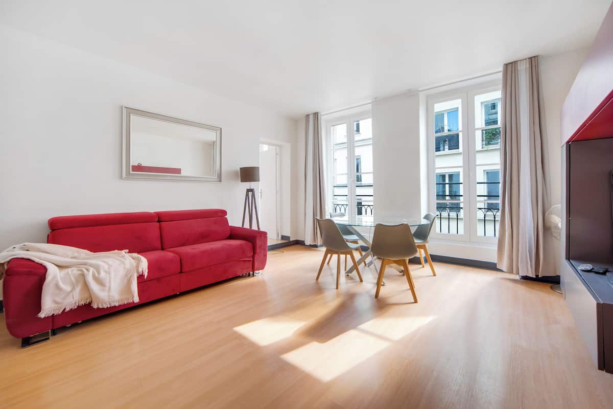 A modern living room with a red sofa, a dining table with chairs, a floor lamp, and large windows offering a view of the outside.