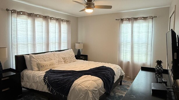 Quarto com cama, escrivaninha, cadeira, ventoinha no teto e janela com cortinas.