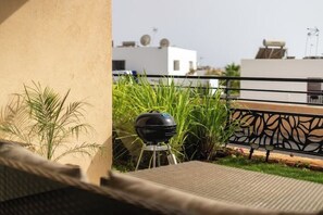 En balkon med grill, plantekasser og udsigt til bygninger i baggrunden.